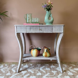 May include: A pale wooden side table with a drawer and a lower shelf. It displays a green jug and glasses, a fish-shaped vase with flowers, and two ceramic pitchers. The table rests on a patterned rug. The table is approximately 76 cm high.