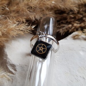 Op de afbeelding: Een zilveren ring met een zwarte vierkante bedel met een gouden wielontwerp.