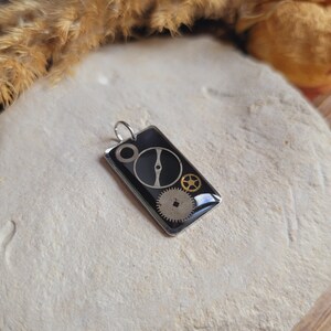 May include: A silver pendant with a black background and gold gears. The pendant is rectangular and has a silver bail.