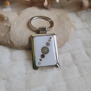 Può includere: Un portachiavi rettangolare argentato con sfondo bianco, incastonato con vari piccoli ingranaggi metallici di orologio. Il portachiavi ha un bordo argentato e un anello argentato nella parte superiore. Gli ingranaggi sono disposti verticalmente.