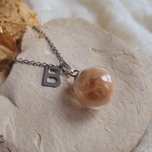 Può includere: Una collana a catena d'argento con un ciondolo a forma di sfera di vetro contenente un ciuffo di capelli castano chiaro. La collana presenta anche un ciondolo d'argento a forma della lettera "B".