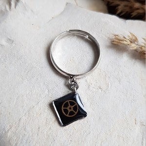 Op de afbeelding: Een zilveren ring met een zwarte vierkante bedel met een gouden tandwielontwerp.