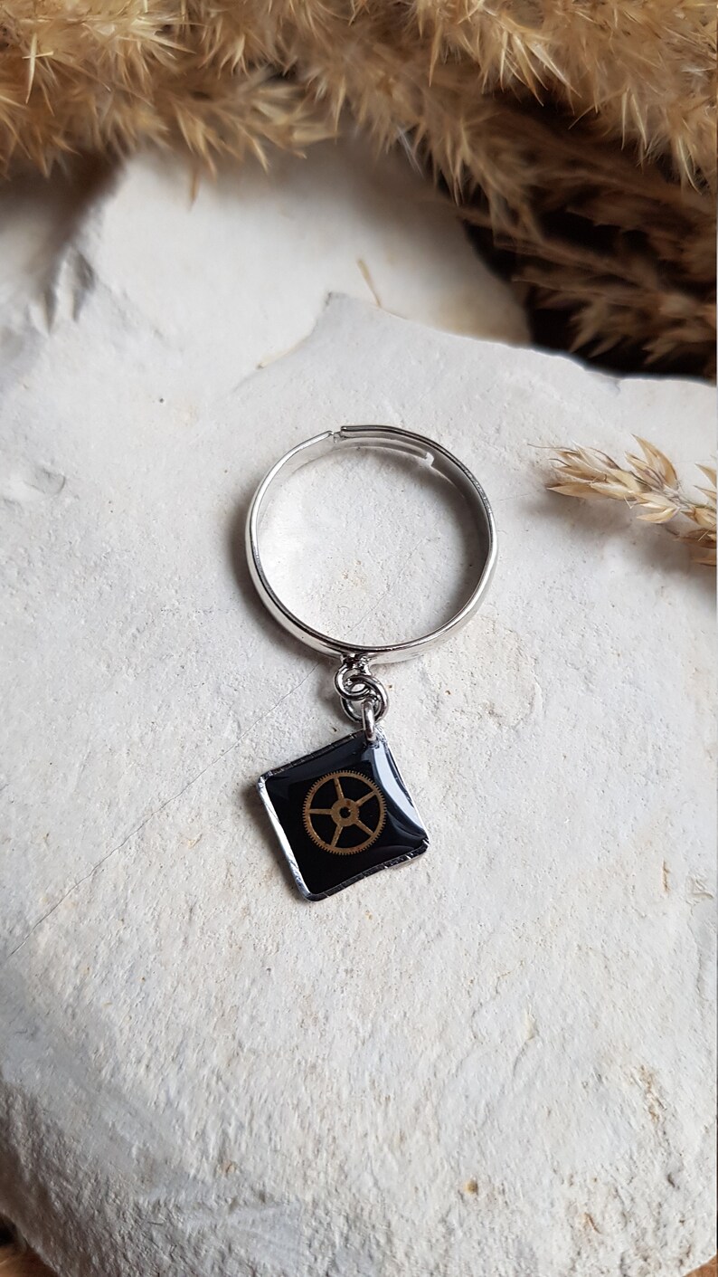 Op de afbeelding: Een zilveren ring met een zwarte vierkante bedel met een gouden tandwielontwerp.