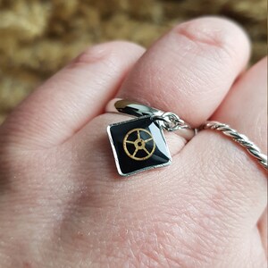 Op de afbeelding: Een zilveren ring met een zwarte vierkante bedel met een gouden tandwielontwerp.