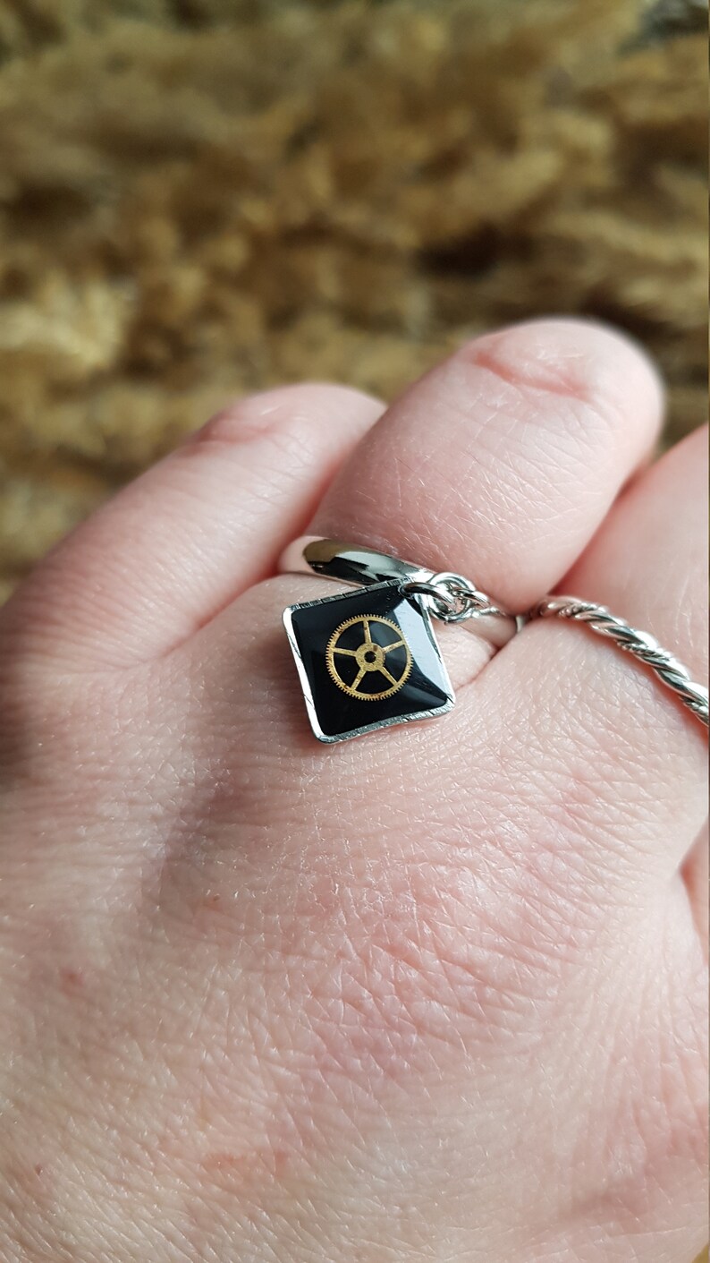 Op de afbeelding: Een zilveren ring met een zwarte vierkante bedel met een gouden tandwielontwerp.