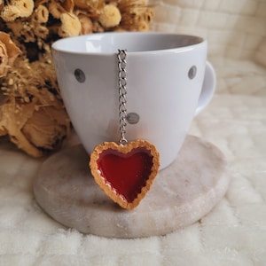 May include: A heart-shaped tea infuser with a red jelly filling, attached to a silver chain. The infuser is hanging from the edge of a white teacup with black polka dots.