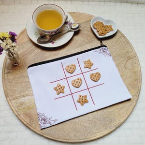 May include: A white zippered pouch with a tic-tac-toe grid and heart and star-shaped cookies. A teacup with tea, a small dish of cookies, and a vase of flowers sit on a wooden tray. The pouch has a floral design.