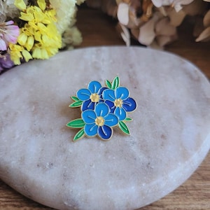 Puede incluir: Un broche de flores de esmalte azul con ribetes dorados y hojas verdes. El broche presenta un grupo de flores de nomeolvides con centros amarillos. El broche está sobre una superficie de piedra clara.