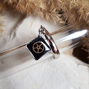 Op de afbeelding: Een zilveren ring met een zwarte vierkante bedel met een gouden tandwielontwerp. De bedel is met een zilveren ketting aan de ring bevestigd.