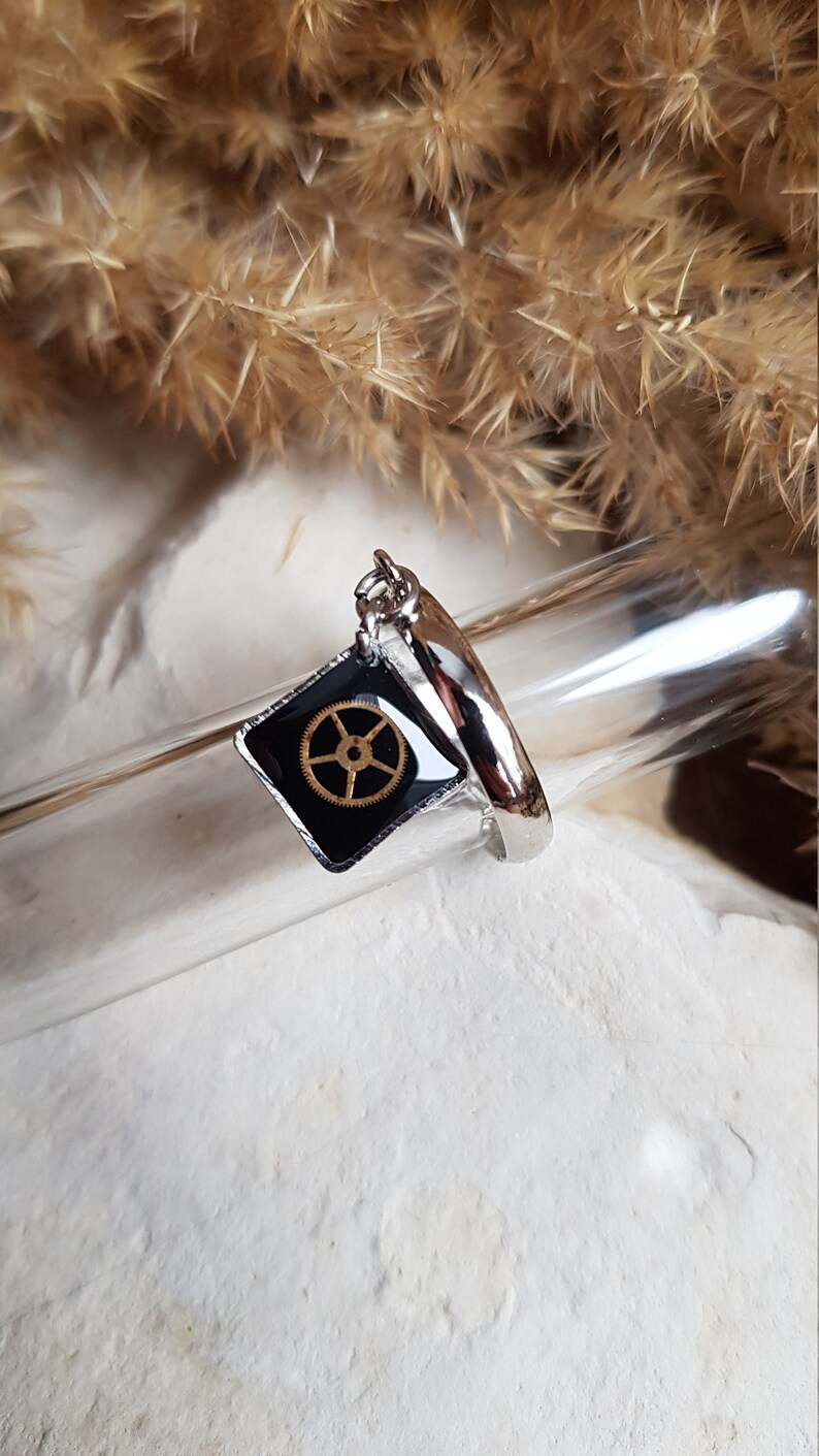 Op de afbeelding: Een zilveren ring met een zwarte vierkante bedel met een gouden tandwielontwerp. De bedel is met een zilveren ketting aan de ring bevestigd.
