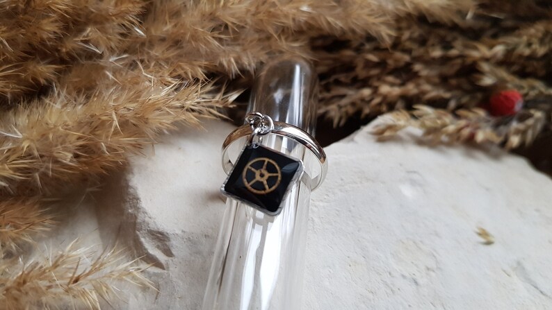 Op de afbeelding: Een zilveren ring met een zwarte vierkante bedel met een gouden wielontwerp.