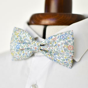 Peut inclure: Un nœud papillon à motifs floraux bleus, jaunes et roses, posé sur une chemise blanche. Le nœud est noué et placé sur un mannequin de cou en bois, mettant en valeur le motif complexe du tissu.