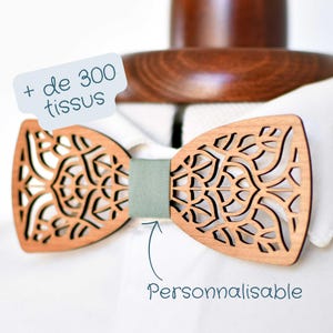 Può includere: Un papillon in legno con un design delicato e intricato tagliato al laser. Il papillon è di colore marrone chiaro e ha un nastro in tessuto verde. Il testo "de 300 tissus" e "Personnalisable" è visibile nell'immagine.