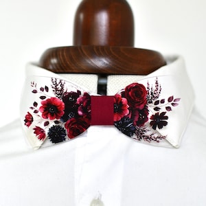 Peut inclure: Un nœud papillon en acrylique transparent orné d'un motif floral imprimé de roses, d'anémones et de feuillages dans des tons rouge profond et violet foncé. Le centre est entouré d'une bande de tissu rouge foncé sur une chemise blanche.