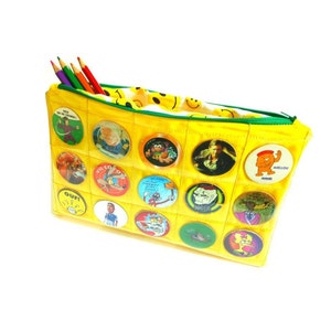 Peut inclure: Une trousse jaune avec une fermeture éclair verte, remplie de crayons de couleur. La trousse est décorée de boutons de personnages de dessins animés. L'intérieur est doublé d'un motif de visages souriants jaunes.