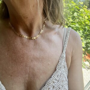 Puede incluir: Un collar dorado con un diseño de hojas. El collar se lleva alrededor del cuello y es el punto focal de la imagen. El fondo está borroso, con follaje verde visible.