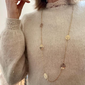 Peut inclure: Un long collier en chaîne dorée avec de délicats pendentifs en forme de feuilles ajourées. Le collier est porté par une personne portant un pull crème.