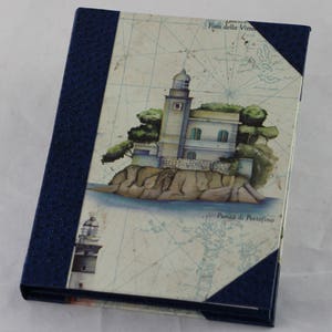 Peut inclure: Un journal relié en cuir bleu sur le thème nautique. La couverture représente un phare sur une petite île avec un fond de carte. Le texte sur la couverture indique "Faro della Vitto" et "Punta di Portofino".