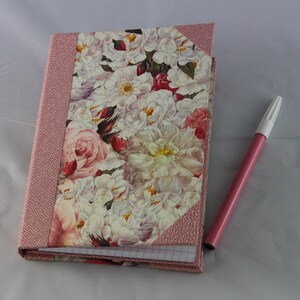 Peut inclure: Un carnet rose avec une couverture à motif floral. La couverture présente une variété de fleurs blanches, roses et rouges. Un stylo rose est posé à côté du carnet.