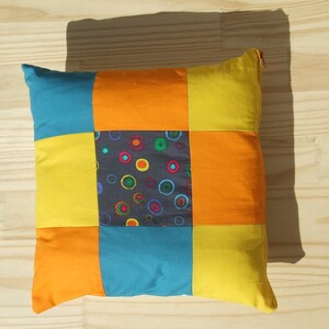 Peut inclure: Un coussin patchwork avec un motif géométrique coloré. Le coussin est composé de six carrés, dont deux sont jaunes, deux sont orange, un est bleu et un est gris avec des cercles colorés.