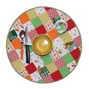 Peut inclure: Un dessous de plat rond en patchwork avec un motif rouge, vert, orange et blanc. Le dessous de plat a un bol, une cuillère, un couteau et un verre dessus.