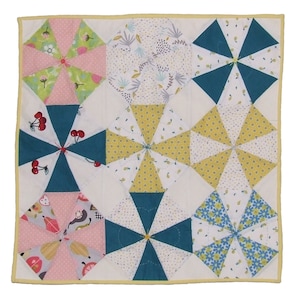 Peut inclure: Une courtepointe avec un fond blanc et neuf carrés colorés. Chaque carré présente un motif différent de tissu turquoise, jaune, rose et blanc. La courtepointe est bordée d'une bordure jaune.