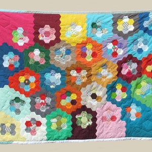 Peut inclure: Une courtepointe colorée composée de patchs de tissu hexagonaux dans diverses couleurs et motifs. La courtepointe est composée de nombreux tissus différents, y compris des imprimés floraux, des motifs géométriques et des couleurs unies. La courtepointe est bordée d'une bordure blanche.