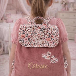 Pode incluir: Uma mochila de veludo rosa seco com uma aba de estampa floral, duas aplicações de corações florais e o nome Céleste bordado em fio dourado. A mochila inclui alças a condizer com estampa floral e um elástico de cabelo coordenado.