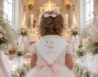 May include: A white triangular bib with lace trim featuring pink embroidery that reads Baptême Maelya and 25 juillet 2026, worn over a white dress with lace sleeves and a pink satin waist ribbon, complemented by a small pink hair bow and flowers.