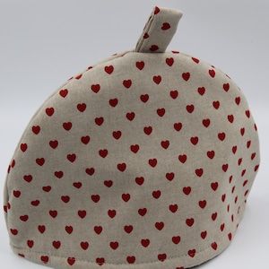 Puede incluir: Un calienta-té beige con un patrón de corazones rojos. El calienta-té tiene un asa de bucle en la parte superior.