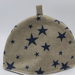 Puede incluir: Un calienta-tetera beige con un patrón de estrellas azul marino. El calienta-tetera tiene un lazo en la parte superior para colgarlo.