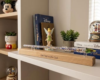 May include: A clear glass wand filled with blue and gold glitter, resting on a rectangular wooden stand engraved with the text Faith Trust and Pixie Dust. The wand features a twisted glass handle and a green tip, displayed on a shelf.