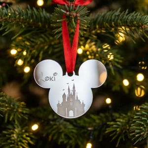 Op de afbeelding: Een zilverkleurig ornament met spiegelafwerking in de vorm van een muizenkop-silhouet. Het ornament heeft een gegraveerde kasteelafbeelding in het midden en de naam Loki op het linkeroor. Het hangt aan een felrood lint gestrikt tot een strik.