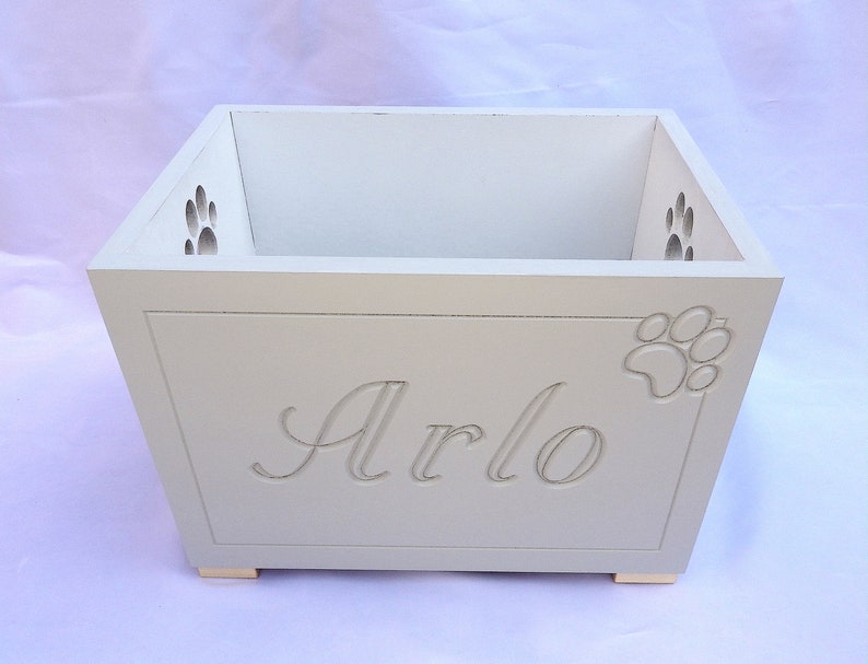 Op de afbeelding: Een rechthoekige witte houten opbergdoos met de naam Arlo in cursief schrift gegraveerd op het voorpaneel. De doos is voorzien van uitgesneden pootafdrukken op de rechtervoorkant en op de zijpanelen aan de binnenkant, met kleine voetjes.