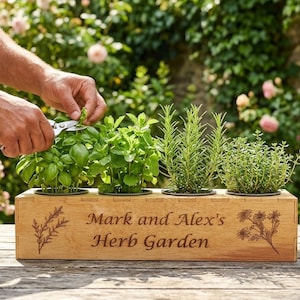 Op de afbeelding: Een rechthoekige, lichtbruine houten plantenbak met vier kleine kruiden in potten. De voorkant van de bak is gegraveerd met de tekst "Mark and Alex's Herb Garden", geflankeerd door botanische illustraties van takjes en bloemen.
