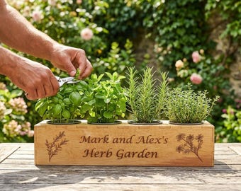 May include: A rectangular light brown wooden planter box containing four small potted herbs. The front of the box is engraved with the text "Mark and Alex's Herb Garden" flanked by botanical illustrations of sprigs and flowers.