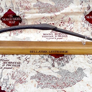 May include: A black wand with intricate details rests on a wooden stand. The stand is engraved with the words "Bellatrix Lestrange" and "Messrs Moony Wormtail Padfoot & Prongs are proud to present".