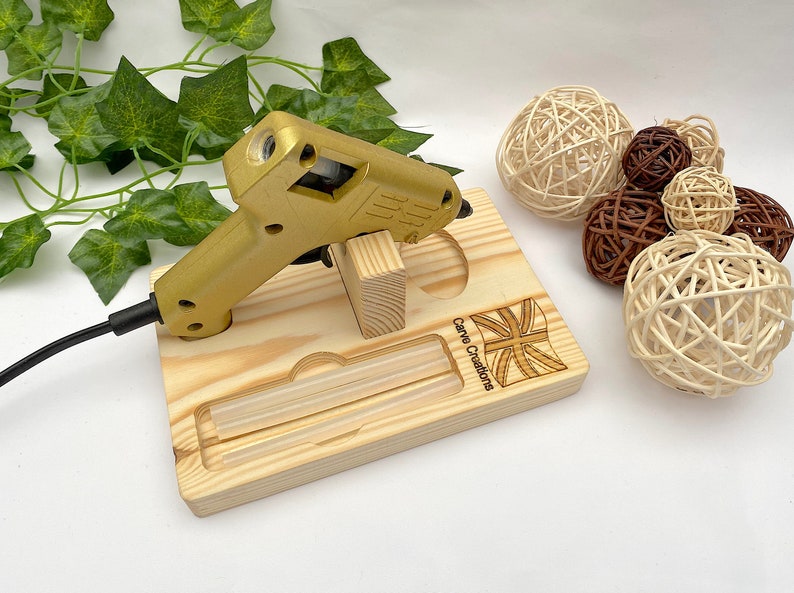 Puede incluir: Una pistola de pegamento de color dorado sobre un soporte de madera clara con un compartimento integrado para dos barras de pegamento. La base de madera presenta un dise&ntilde;o grabado de la bandera Union Jack y el texto Carva Creations, con cable negro.