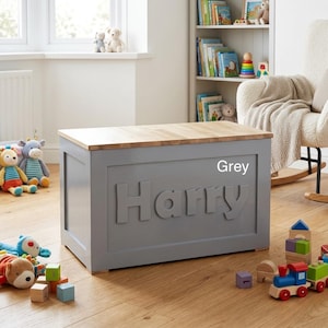 May include: A rectangular grey wooden toy chest with a natural light wood top. The front panel features the name Harry in large, raised, matching grey letters. The chest has a clean, solid construction with a smooth finish and a flat lid.