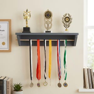 Op de afbeelding: Een donkergrijze houten wandplank met een horizontale stang voor het ophangen van medailles. Drie goudkleurige trofeeën staan op de bovenkant. Zes kleurrijke linten met ronde medailles hangen verticaal aan de houten stang.
