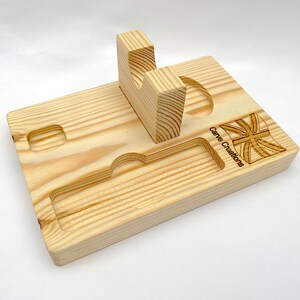 Puede incluir: Una bandeja organizadora rectangular de madera clara que presenta un soporte central vertical con un recorte curvo, una ranura rectangular larga y empotrada, y dos hendiduras circulares m&aacute;s peque&ntilde;as. El texto Carve Creations y una bandera est&aacute;n grabados.
