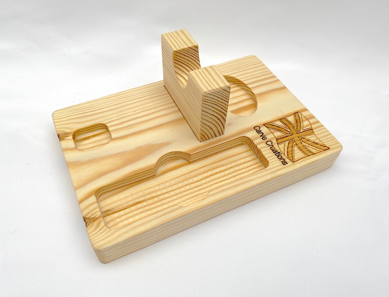Puede incluir: Una bandeja organizadora rectangular de madera clara que presenta un soporte central vertical con un recorte curvo, una ranura rectangular larga y empotrada, y dos hendiduras circulares m&aacute;s peque&ntilde;as. El texto Carve Creations y una bandera est&aacute;n grabados.