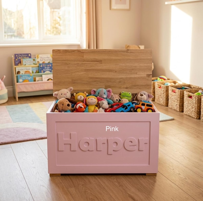 May include: A pink wooden toy chest with a natural wood lid, featuring the name Harper embossed in large letters on the front. The chest is filled with various plush stuffed animals and small toy vehicles, providing storage for children's play items.
