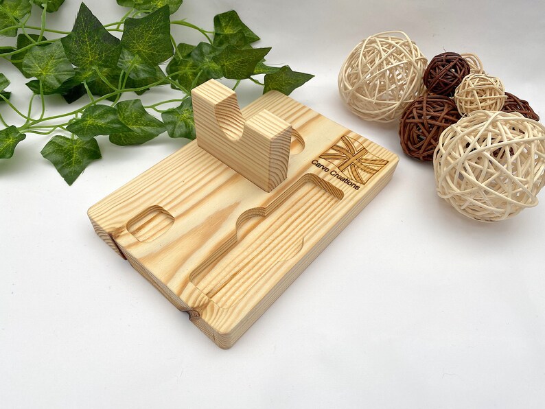 Puede incluir: Un soporte organizador rectangular de madera clara que presenta una pieza de soporte vertical con un recorte en forma de U, una ranura rectangular y un canal largo empotrado. La superficie de madera est&aacute; grabada con una bandera Union Jack y el texto Carve Creations.