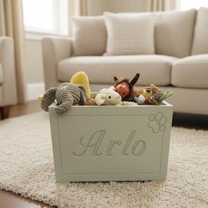 Op de afbeelding: Een licht saliegroene houten speelgoedkist met de naam Arlo in cursief schrift gegraveerd op het voorpaneel, vergezeld van een pootafdruk-icoon. De kist is gevuld met diverse pluche knuffeldieren, waaronder een olifant en een schaap.