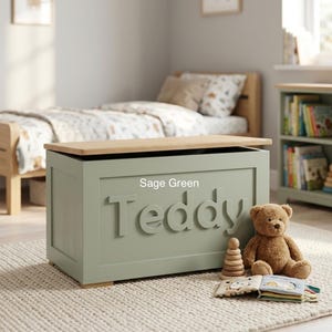 May include: A sage green wooden toy chest with a natural wood-toned lid. The front of the chest features the name Teddy in raised, sage green lettering. A brown plush teddy bear and a small open book are positioned in front of the chest.