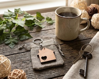 May include: A wooden house-shaped keychain with a heart-shaped cutout, attached to a silver metal ring. The keychain rests on a small, light brown burlap drawstring pouch, accompanied by a metal key and a ceramic mug filled with dark liquid.