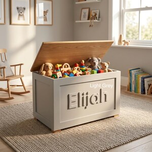 May include: A light grey wooden toy chest with a natural wood-toned lid. The front panel features the name Elijah in raised lettering. The chest is filled with various colorful wooden blocks and plush stuffed animals, including a bear and a rabbit.