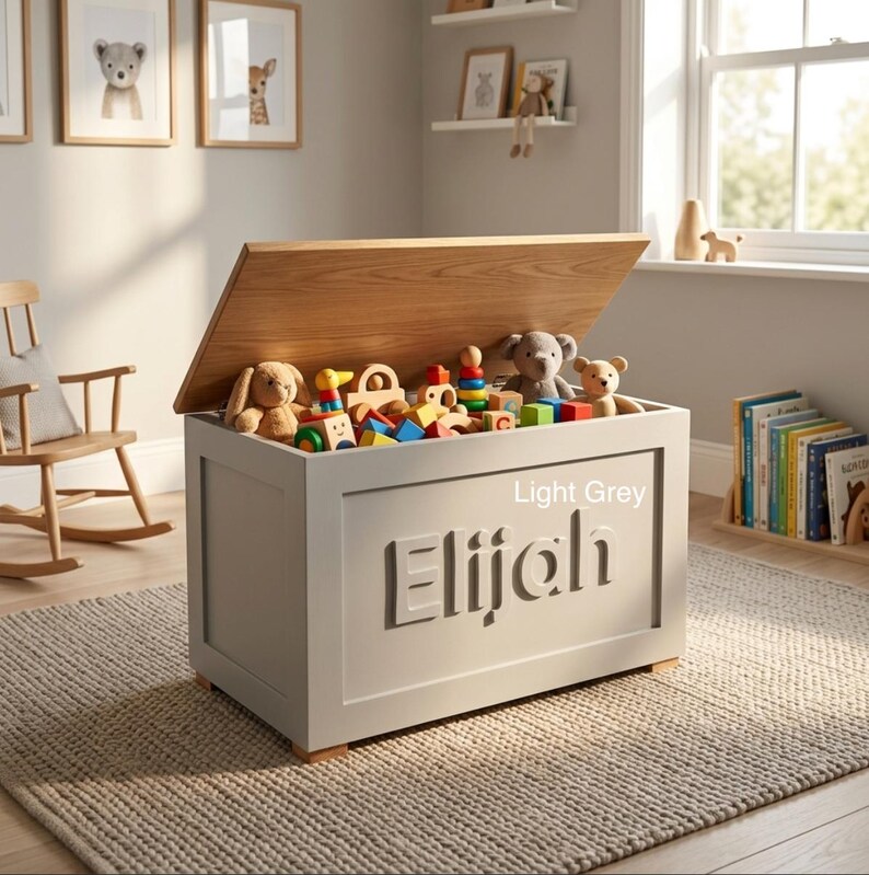 May include: A light grey wooden toy chest with a natural wood-toned lid. The front panel features the name Elijah in raised lettering. The chest is filled with various colorful wooden blocks and plush stuffed animals, including a bear and a rabbit.