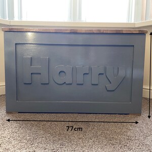 May include: A rectangular wooden toy chest painted in a matte gray finish with the name Harry in raised lettering on the front panel. The chest features a natural wood top lid and measures 77 cm in width by 48 cm in height as indicated by arrows.